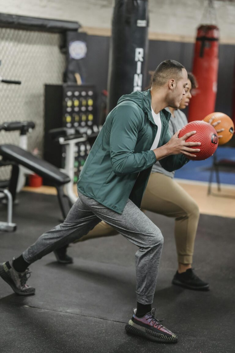 Side view of sporty young multiethnic male trainer and female client doing forward lunges exercise with medicine ball during workout in gym together